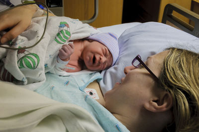 High angle view of mother looking at crying newborn son while lying on bed in hospital