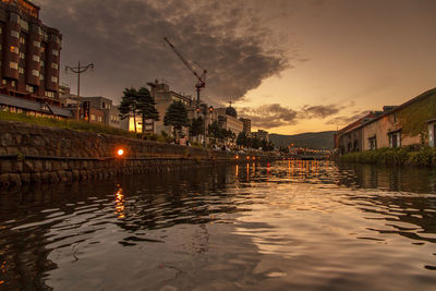 River by illuminated buildings against sky at sunset