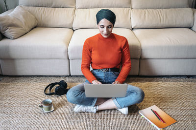 High angle of glad muslim woman in casual clothes sitting cross legged on floor near sofa and browsing data on netbook while working on remote project at home