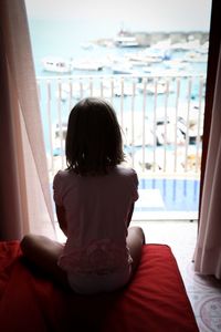 Rear view of girl sitting on bed while looking at sailboats through window