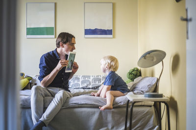 Man showing photograph on digital tablet to son sitting in bedroom at apartment