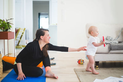 Side view of working mother reaching for daughter while sitting on exercise mat in living room at home