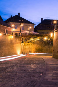 Light trails on road by buildings against sky at night