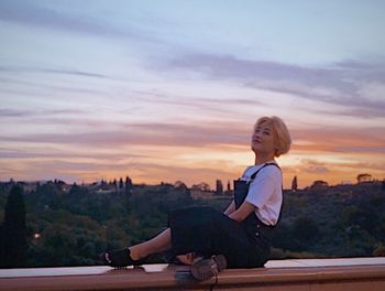Side view of woman sitting against sky during sunset