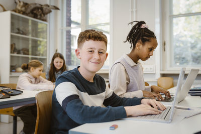 Portrait of smiling schoolboy with laptop by female friend in classroom