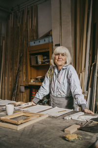 Smiling senior female carpenter leaning near workbench at repair shop