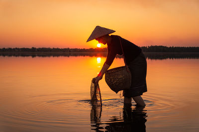 Woman standing by lake against sky during sunset
