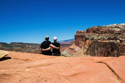 Couple with arm around sitting on rock against clear sky