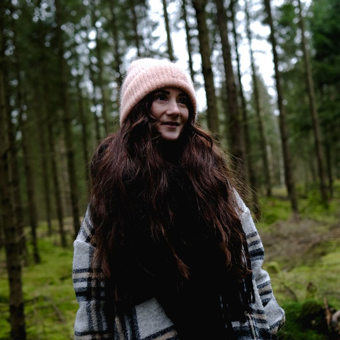 forest, tree, long hair, one person, land, plant, adult, clothing, woodland, hairstyle, nature, leisure activity, portrait, women, young adult, smiling, warm clothing, lifestyles, hat, brown hair, front view, standing, day, looking at camera, waist up, knit hat, pine woodland, pinaceae, focus on foreground, coniferous tree, happiness, outdoors, winter, non-urban scene, natural environment, casual clothing, wilderness, pine tree, tree trunk, trunk, three quarter length, autumn, beanie, beauty in nature, emotion, person, looking
