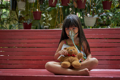 Girl wearing oxygen mask while sitting with teddy bear on bench