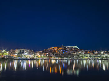 Illuminated buildings by sea against clear blue sky at night