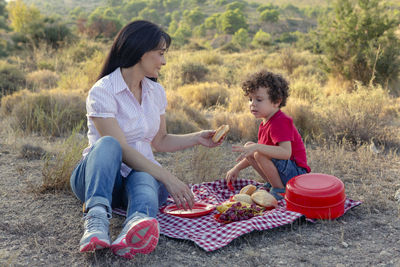 Mom offering her son a sandwich on a hike in the bush for a picnic on a red and whitecheckered 