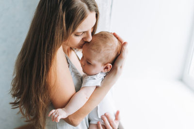 Young woman mom with long hair holding baby girl on hands near window at home