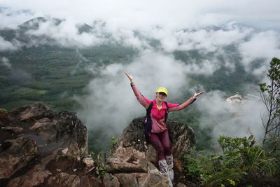 High angle view of female hiker sitting on rock with arms outstretched at mountain peak