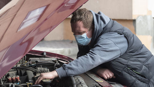 Mechanic in a protective medical mask checks a car engine during a covid-19 coronavirus pandemic