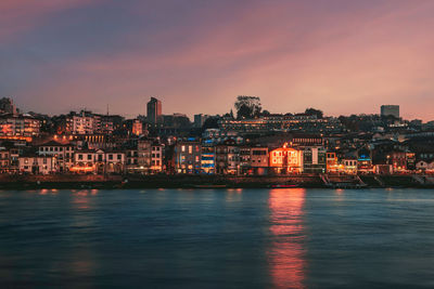 Illuminated buildings by river against sky at sunset