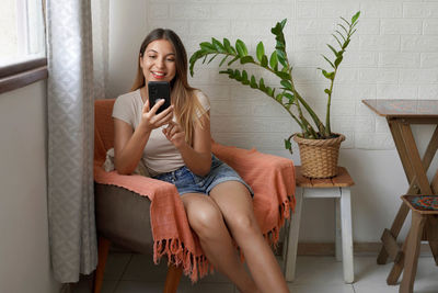 Happy young latin woman sitting on armchair holding mobile phone doing ecommerce shopping