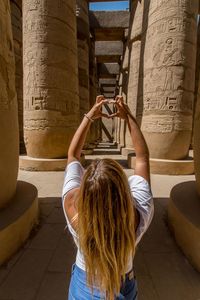 Rear view of woman making hand heart shape while standing amidst architectural column