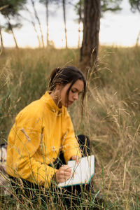 Peaceful thoughtful female sitting with crossed legs in dry meadow and writing thoughts in diary while relaxing at weekend in countryside