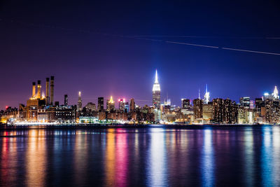 Illuminated buildings in city by river against clear sky at night