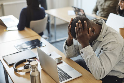 Tired male student with head in hands sitting by laptop on desk in classroom