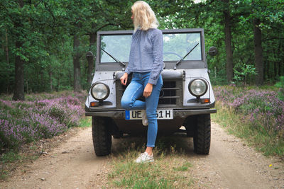 Full length of woman standing by car on dirt road amidst forest