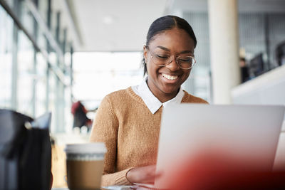 Smiling young female student using laptop while studying at table in university cafeteria