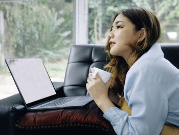 Smiling young woman holding coffee cup by laptop while sitting on sofa