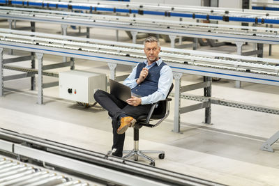 Businessman with laptop sitting amidst conveyor belt at factory