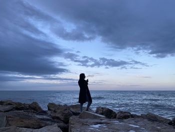 Woman standing on rock looking at sea shore against sky