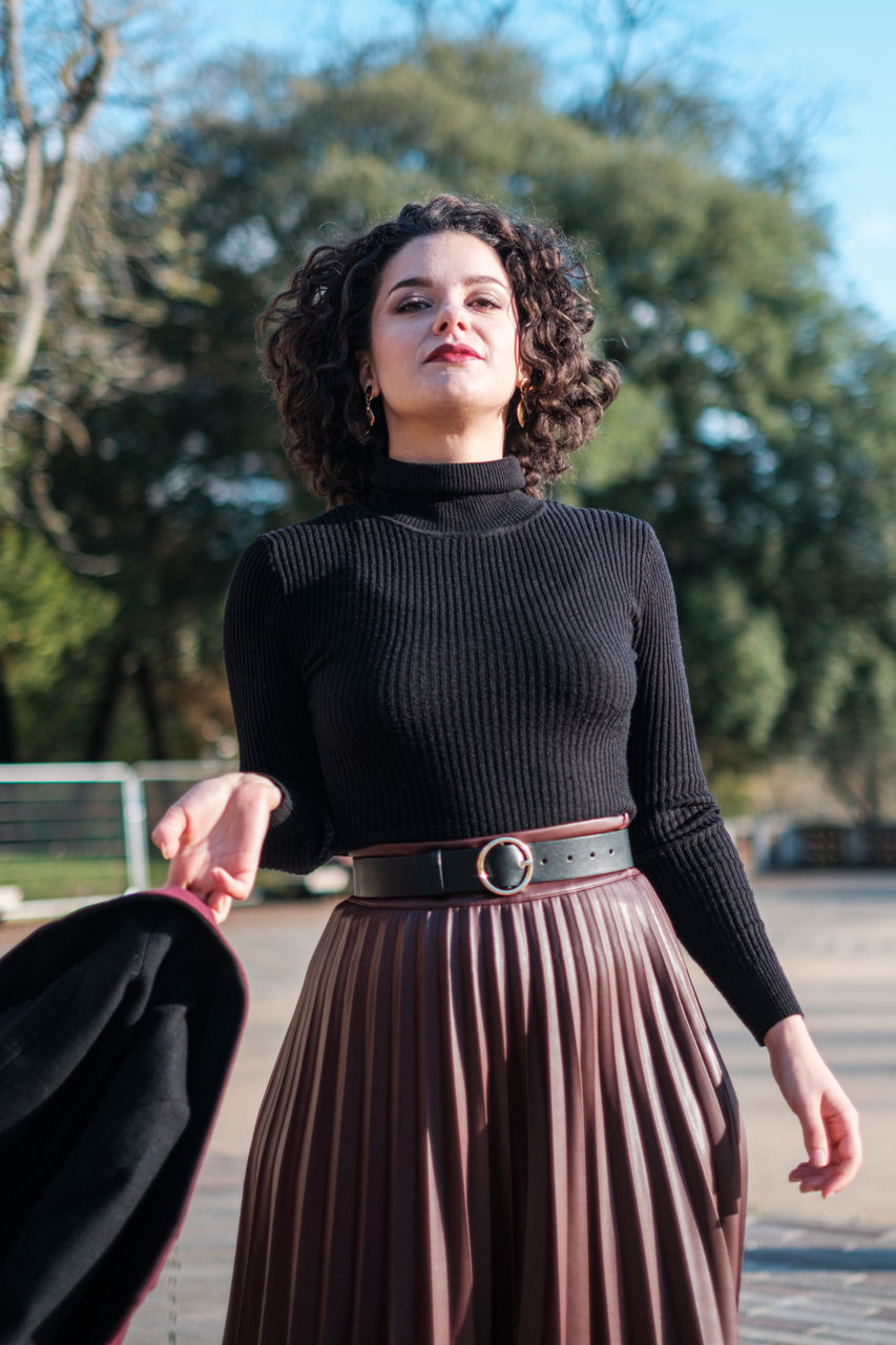 skirt, women, one person, adult, fashion, dress, clothing, spring, hairstyle, portrait, standing, photo shoot, young adult, female, brown hair, curly hair, long hair, nature, tree, emotion, three quarter length, person, looking at camera, black, lifestyles, looking, front view, day, outdoors, pattern, elegance, focus on foreground, smiling, arts culture and entertainment, miniskirt