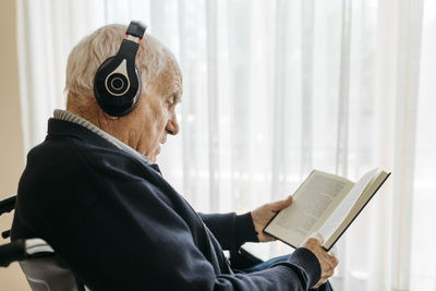 Senior man sitting in wheelchair reading a book while listening music with headphones