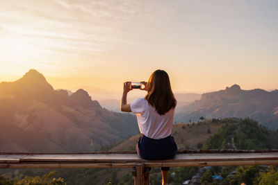 Rear view of woman photographing against sky during sunset