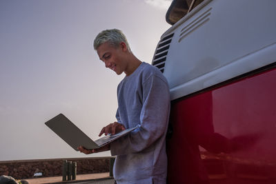 Low angle view of smiling boy using laptop standing by car against sky