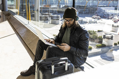 Stylish bearded guy in knitted hat and eyeglasses sending using phone while sitting with laptop against glass wall in airport