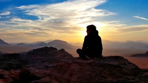 Silhouette teenage boy sitting on mountain against cloudy sky