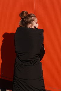 Fashionable teenage girl wearing black warm clothing standing against red wall during sunny day