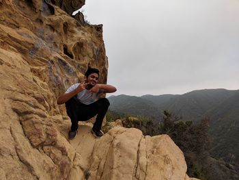 Full length of young man sitting on rock while gesturing against mountains