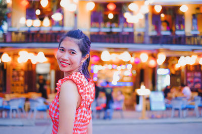 Portrait of smiling woman standing against illuminated restaurant