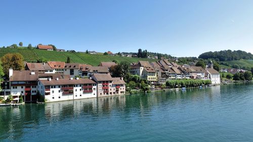 Houses by river and buildings against clear blue sky