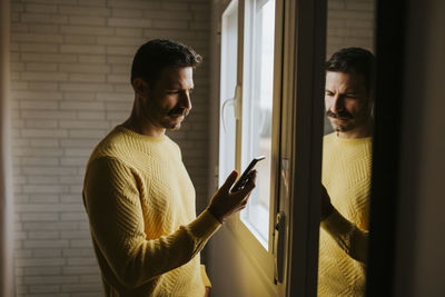 Serious man using smart phone while standing by window at home