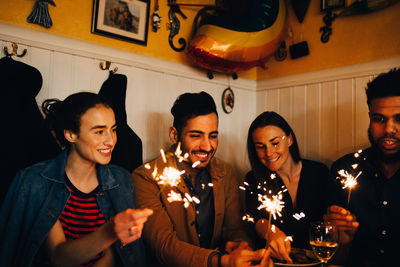 Happy young multi-ethnic male and female friends holding burning sparklers in restaurant during dinner party