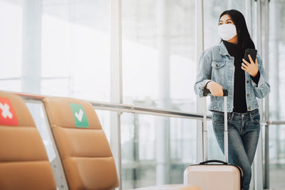Woman wearing mask holding smart phone while standing by window at airport
