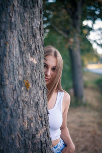Portrait of young woman looking away while hiding behind tree trunk