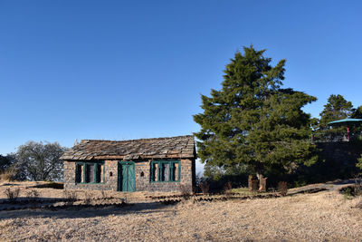 Trees and abandoned house on field against clear blue sky