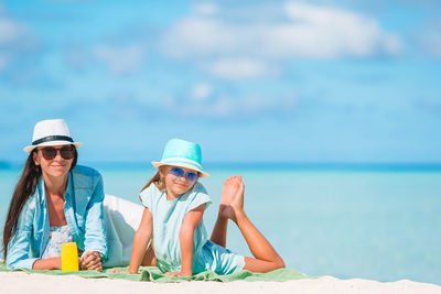 Mother and daughter wearing hat lying down on beach against sky