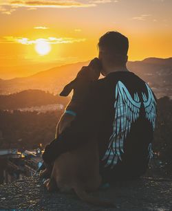 Rear view of man and dog sitting against sky during sunset