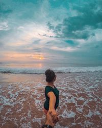 Couple holding hands while standing on shore against sky during sunset