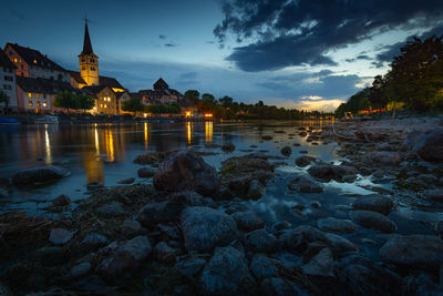 Illuminated buildings by river against sky at dusk