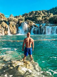 Full length of shirtless man standing on rocks against blue sky